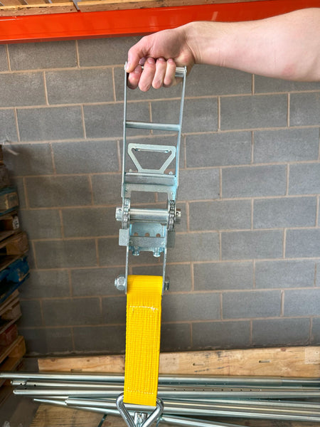 Hand holding a 10,000kg Ratchet Strap with Claw Hooks, featuring a yellow handle and metal components, suitable for securing large industrial machinery.